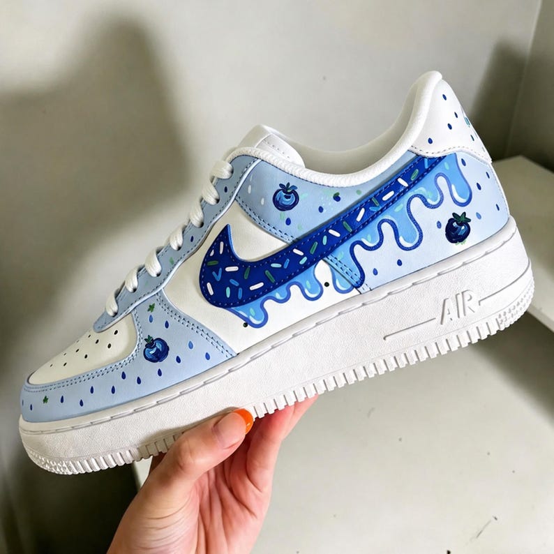 May include: White and light blue Nike Air Force 1 sneakers with a custom blueberry design. The sneakers feature a white sole, light blue accents, and a dark blue Nike swoosh. The design includes blueberry illustrations and dripping paint details.