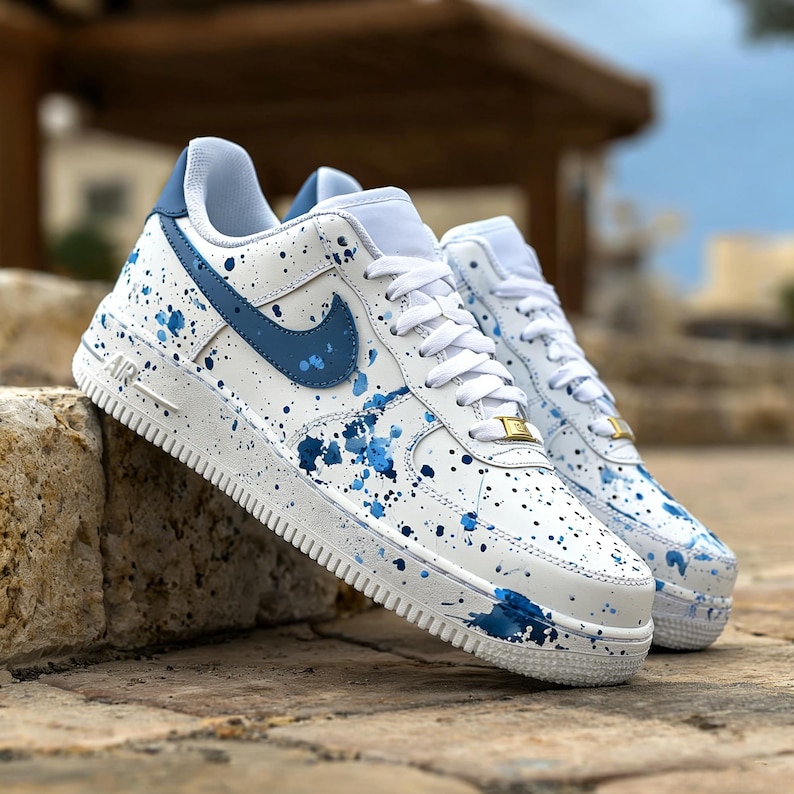 May include: White sneakers with blue accents and a splatter paint design. The shoes feature a blue Nike swoosh, laces, and a gold detail on the laces. The soles are white with a textured pattern.