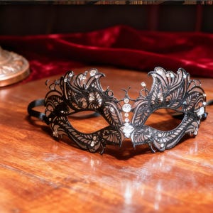 Op de afbeelding: Een zwart masker voor een gemaskerd bal met ingewikkelde filigraan details en sprankelende strass-steentjes. Het masker heeft een symmetrisch ontwerp en wordt vastgezet met een satijnen lint. Perfect voor een kostuumfeest.