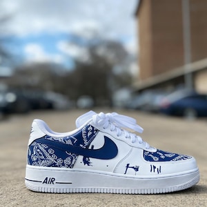 May include: White and blue Air Force 1 sneakers with a custom design. The sneakers feature a white base with blue accents, including a bandana-style pattern on the heel and toe, and a blue Nike swoosh. The word "AIR" is printed on the white midsole.