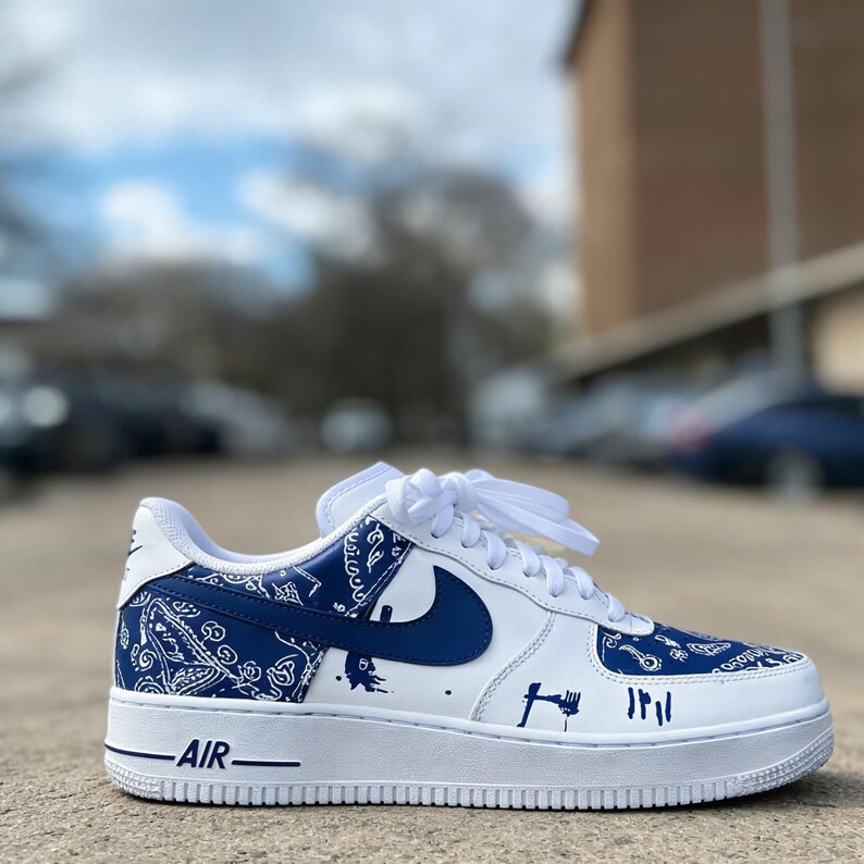 May include: White and blue Air Force 1 sneakers with a custom design. The sneakers feature a white base with blue accents, including a bandana-style pattern on the heel and toe, and a blue Nike swoosh. The word "AIR" is printed on the white midsole.