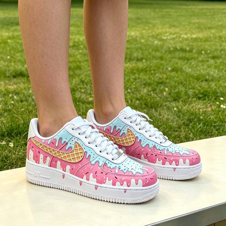 May include: White sneakers with a custom ice cream design. The shoes feature a pink and light blue color scheme with a waffle cone design on the side. The soles are white, and the laces are white.