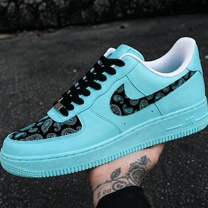 May include: A turquoise sneaker featuring black laces and a black paisley pattern on the toe box and side swoosh. The shoe has a white interior lining, a matching turquoise midsole and outsole, and a silver lace dubrae on the front.