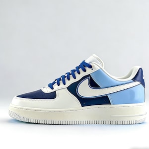 May include: A white, navy blue, and light blue Air Force 1 sneaker. The shoe features a white swoosh, blue laces, and a cream-colored sole. The heel and toe box are navy blue.