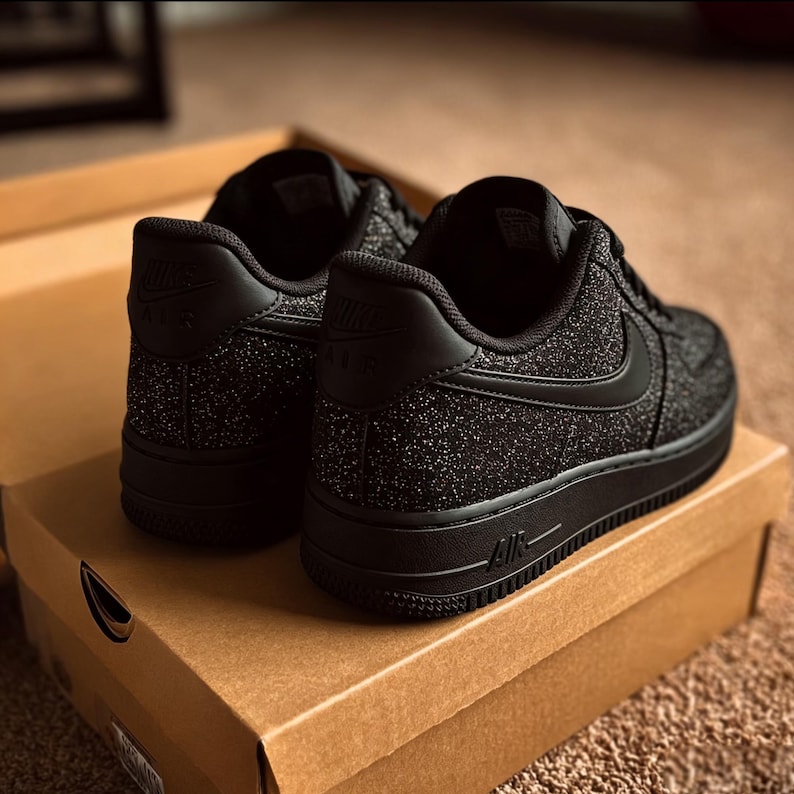 May include: Pair of black Nike Air Force 1 sneakers with a glittery texture, resting on a brown cardboard box. The shoes feature the Nike logo on the heel and side, with the word "AIR" on the sole.