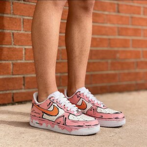 May include: A pair of custom Air Force 1 sneakers in pink, white, and orange. The shoes feature a cartoon-style design with black outlines and the name "Lmara" on the side. The soles are white, and the laces are white.