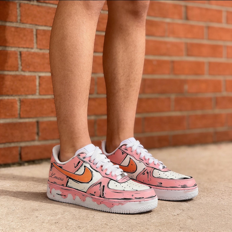 May include: A pair of custom Air Force 1 sneakers in pink, white, and orange. The shoes feature a cartoon-style design with black outlines and the name "Lmara" on the side. The soles are white, and the laces are white.