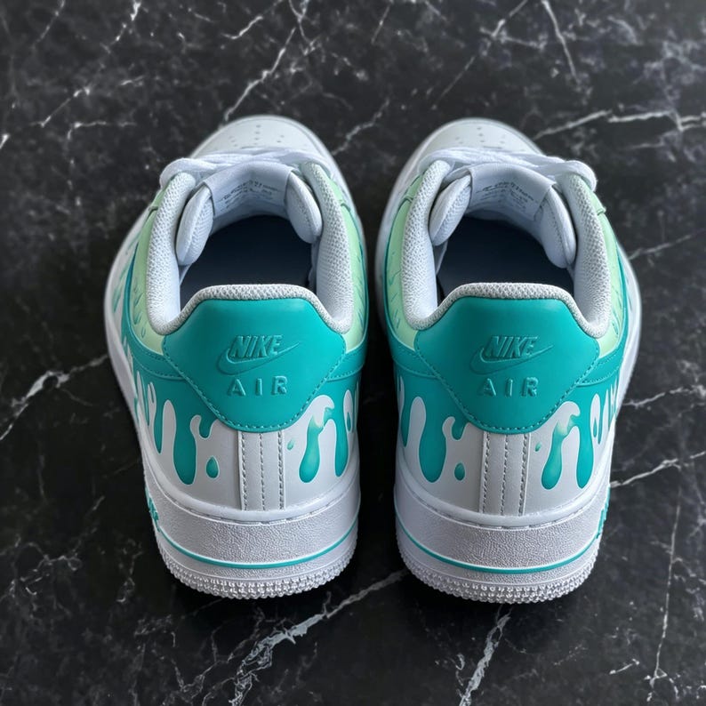 May include: Pair of white Nike Air Force 1 sneakers with teal and light green accents. The teal heel tab features the Nike Air logo, and the design includes a dripping paint effect in teal.