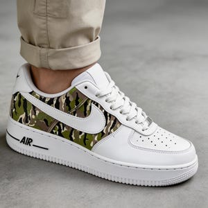 May include: White leather sneaker with a camouflage pattern in shades of green, brown, and black on the side panel. Features a white sole, laces, and the word "AIR" in black. The Nike swoosh is white.
