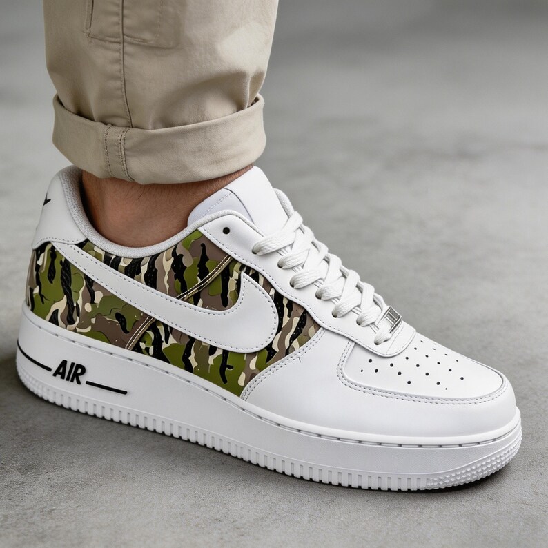 May include: White leather sneaker with a camouflage pattern in shades of green, brown, and black on the side panel. Features a white sole, laces, and the word "AIR" in black. The Nike swoosh is white.