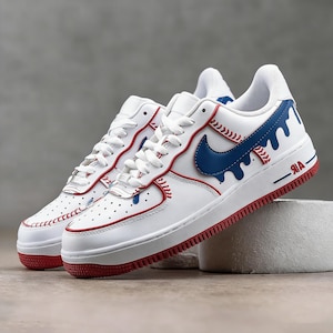 May include: White leather sneakers with red and blue accents. The sneakers feature a red sole, a blue Nike swoosh, and baseball-inspired stitching details. The word "AIR" is printed in red on the side.