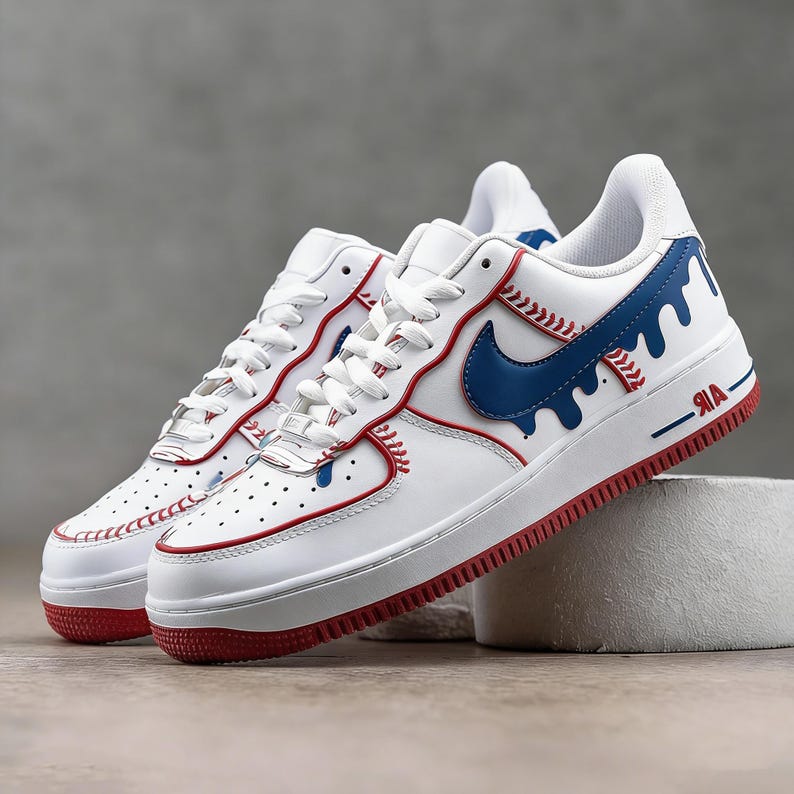 May include: White leather sneakers with red and blue accents. The sneakers feature a red sole, a blue Nike swoosh, and baseball-inspired stitching details. The word "AIR" is printed in red on the side.