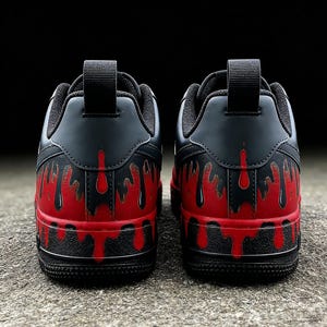May include: Pair of black and red sneakers with a unique design. The shoes feature a black upper with a red, dripping paint effect on the lower portion. The soles are black, and the shoes have a pull tab on the heel.