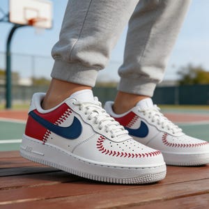Op de afbeelding: Witte leren sneakers met rode en blauwe accenten met honkbalthema. De sneakers hebben een blauwe swoosh, rode stiksels en een witte zool. Het ontwerp is ge&iuml;nspireerd op honkbal, met stikdetails die op een honkbal lijken.