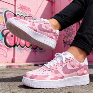 May include: Pink camouflage sneakers with white soles and laces. The sneakers feature a pink Nike swoosh and are set against a pink graffiti background. A stylish and trendy footwear choice.