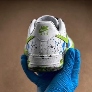 May include: White and green Nike Air Force 1 sneaker with blue and black paint splatter details. The heel features the Nike logo in green. The sole is green and black.