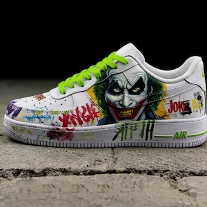 May include: White sneakers with a Joker-themed design. The shoes feature a graphic of the Joker's face, graffiti-style lettering, and green laces. The word "AIR" is visible on the sole.