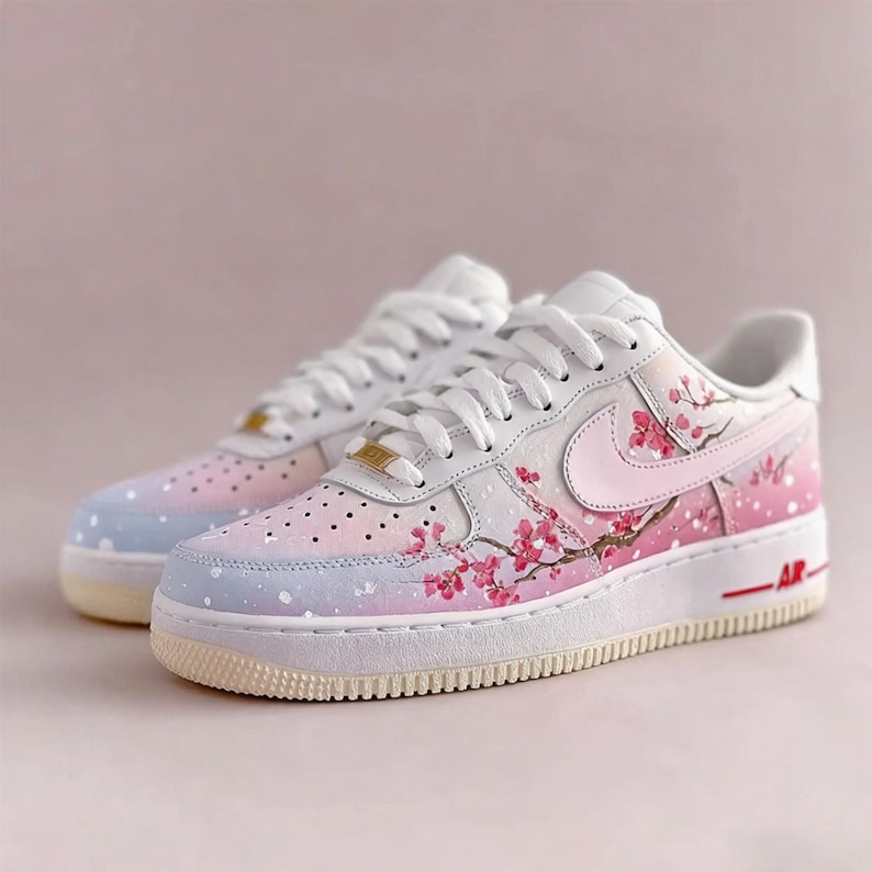 Op de afbeelding: Witte sneakers met een aangepast kersenbloesemdesign. De schoenen hebben een verloop van roze en blauw op de neus en hiel, met roze kersenbloesemtakken en -bloemen geschilderd op de zijkanten. De Nike swoosh is roze.