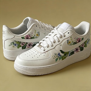 May include: White leather trainers adorned with floral patterns in pink, blue, and yellow. The trainers feature white laces, a white sole, and the Nike logo on the side.