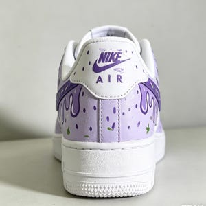 May include: A custom-painted Nike Air Force 1 sneaker. The shoe features a white sole and a light purple upper with a drip design in a darker purple. The heel displays the Nike Air logo.