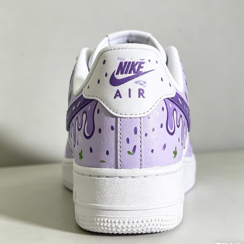 May include: A custom-painted Nike Air Force 1 sneaker. The shoe features a white sole and a light purple upper with a drip design in a darker purple. The heel displays the Nike Air logo.