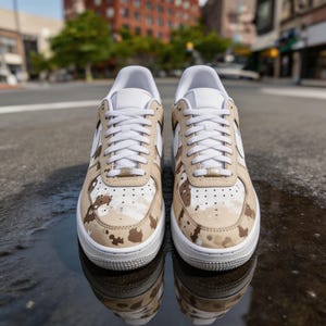 May include: Pair of white and tan sneakers with a camouflage pattern. The shoes have white laces, a white sole, and a tan and brown camouflage design on the upper. The sneakers are sitting on a wet, reflective surface.