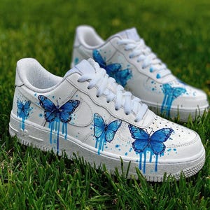 Op de afbeelding: Witte sneakers met blauwe vlinderontwerpen. De schoenen hebben handgeschilderde blauwe vlinders met druipende verfeffecten. De sneakers staan op groen gras.