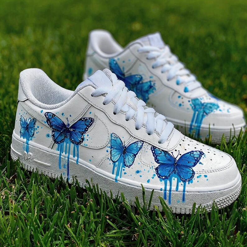 Op de afbeelding: Witte sneakers met blauwe vlinderontwerpen. De schoenen hebben handgeschilderde blauwe vlinders met druipende verfeffecten. De sneakers staan op groen gras.