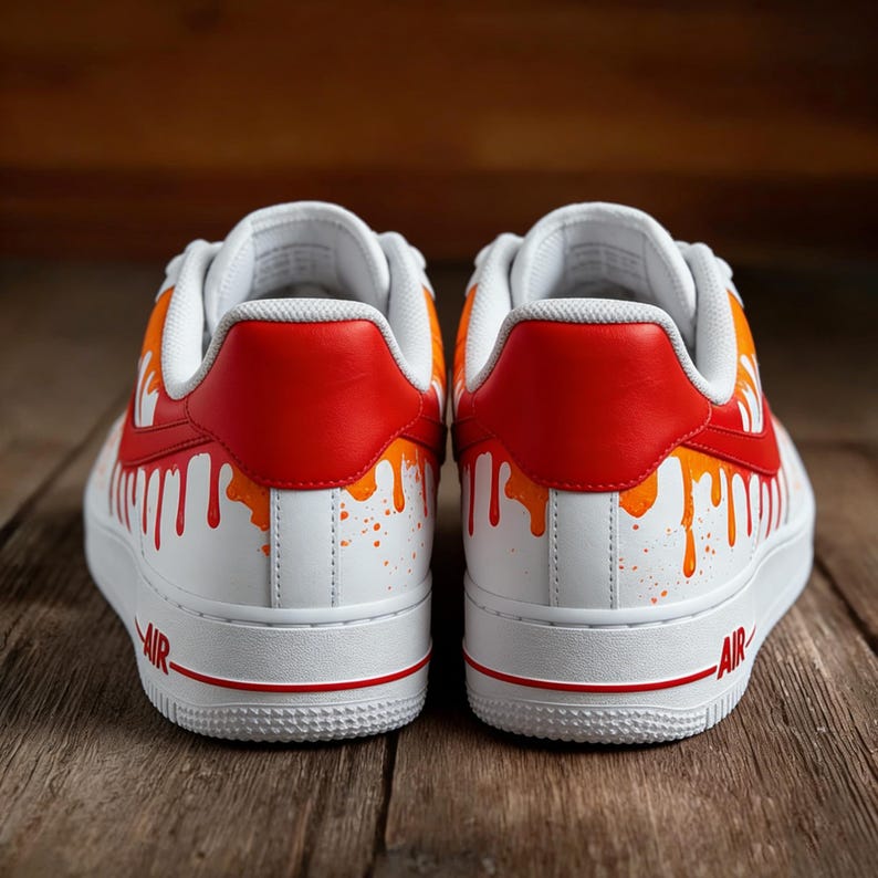 May include: Pair of white sneakers with red and orange paint drip design. The shoes have red heel tabs and orange accents, with the word "AIR" in red on the side. The sneakers are on a wooden surface.