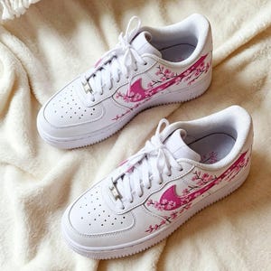 May include: A pair of white sneakers with pink cherry blossom designs. The shoes feature white laces, a pink Nike swoosh, and pink floral accents. The sneakers are on a soft, white surface.