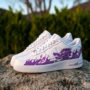 May include: White sneakers with a purple dragon design. The dragon graphic is on the side of the shoe, with flames extending towards the front. The shoes have white laces and a white sole. The word "AIR" is printed on the side of the sole.