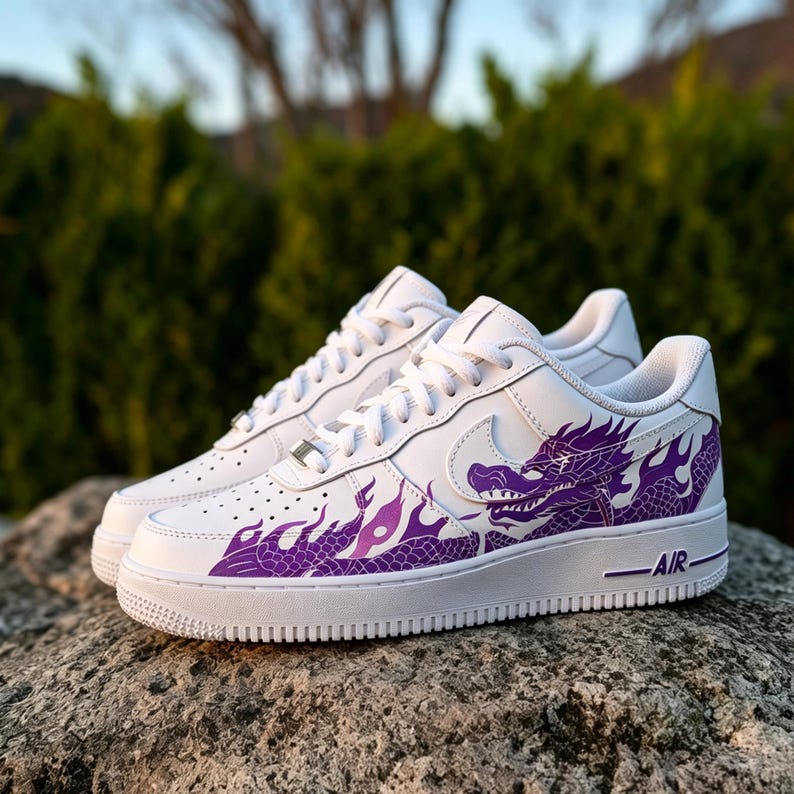 May include: White sneakers with a purple dragon design. The dragon graphic is on the side of the shoe, with flames extending towards the front. The shoes have white laces and a white sole. The word "AIR" is printed on the side of the sole.