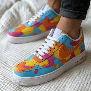 May include: A pair of colorful Nike Air Force 1 sneakers. The shoes feature a vibrant design with blue, orange, yellow, and pink camouflage-style patterns. They have white laces, soles, and the iconic Nike swoosh in orange.