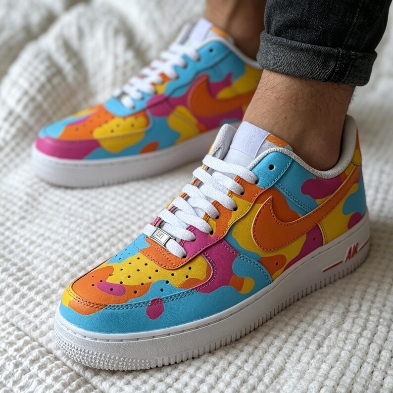 May include: A pair of colorful Nike Air Force 1 sneakers. The shoes feature a vibrant design with blue, orange, yellow, and pink camouflage-style patterns. They have white laces, soles, and the iconic Nike swoosh in orange.