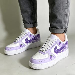 May include: White and light purple sneakers with a grape-themed design. The sneakers feature a white base with light purple accents on the toe and heel. The Nike swoosh is purple with a dripping design, and there are grape illustrations.