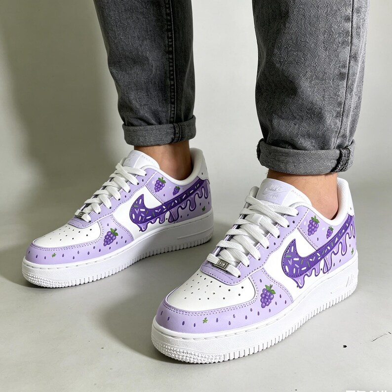 May include: White and light purple sneakers with a grape-themed design. The sneakers feature a white base with light purple accents on the toe and heel. The Nike swoosh is purple with a dripping design, and there are grape illustrations.