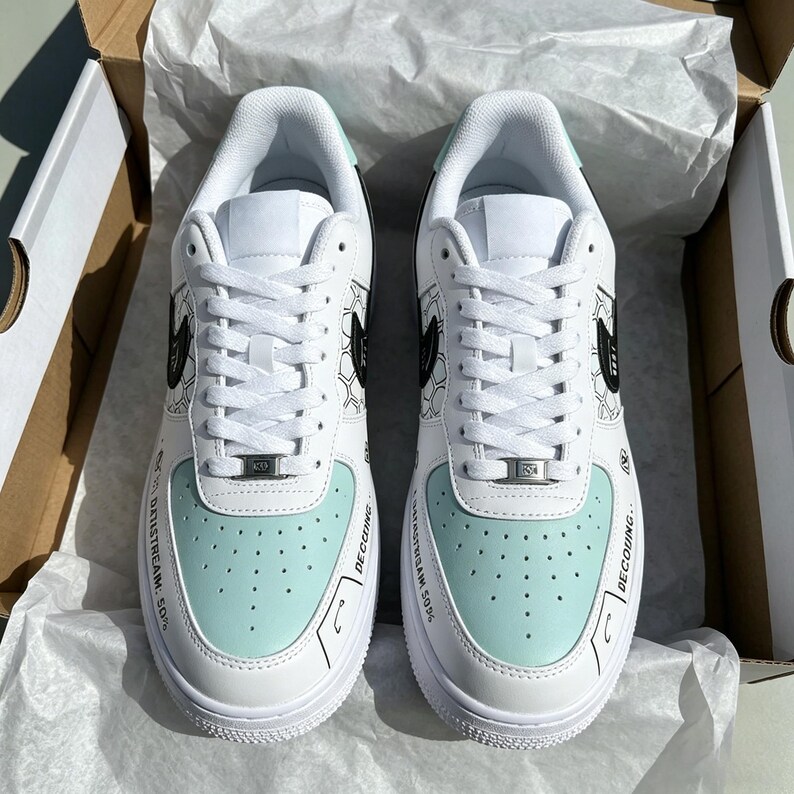 May include: White and light blue sneakers with black accents, displayed in an open cardboard box. The shoes feature a light blue toe box, white laces, and black graphic details. Text is printed on the side of the shoe.