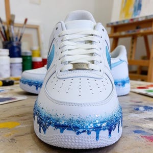 May include: White leather sneakers with blue glitter accents and a dripping paint effect on the soles. The Nike swoosh is light blue with glitter. The laces are white, and the tongue has a silver metal tag.