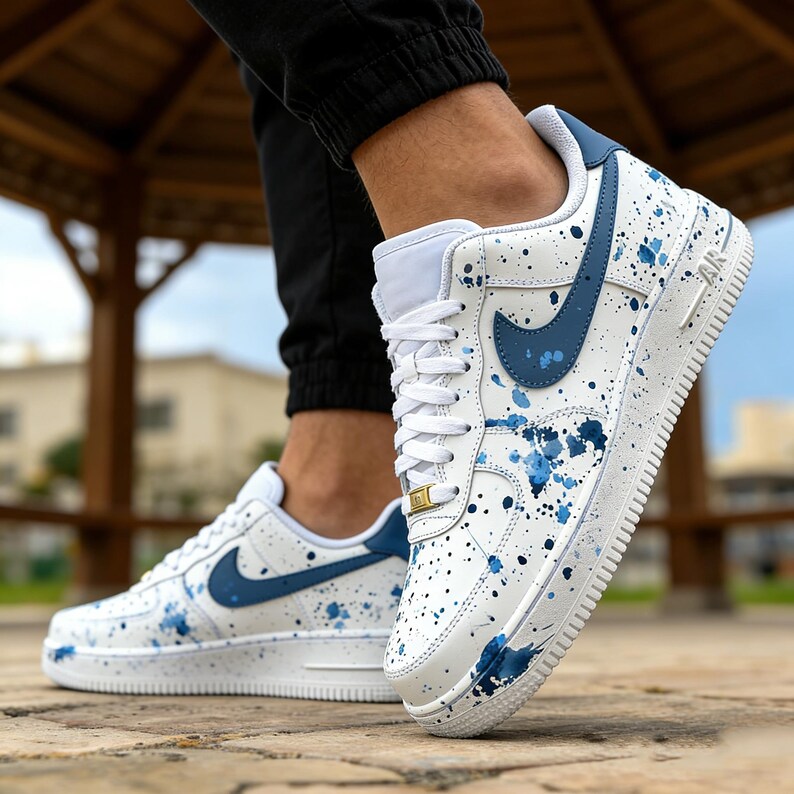 May include: White sneakers with blue accents and paint splatter design. Features a blue Nike swoosh and laces. The shoes are a classic athletic style.