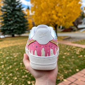 May include: A white Nike Air Force 1 sneaker with a pink donut design. The shoe features pink icing drips, colorful sprinkles, and a white sole. The Nike logo is visible on the heel.