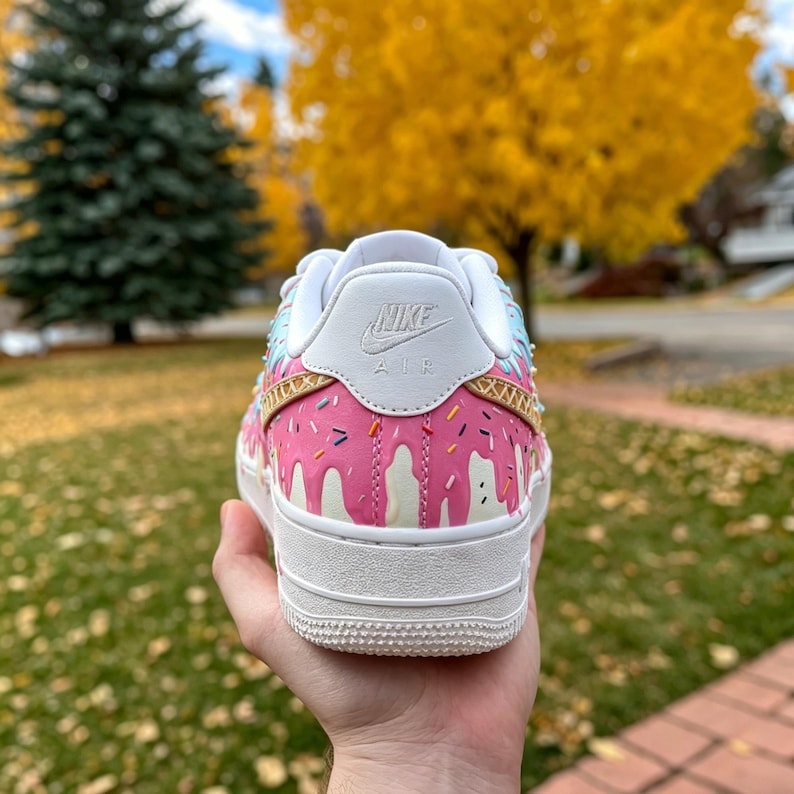 May include: A white Nike Air Force 1 sneaker with a pink donut design. The shoe features pink icing drips, colorful sprinkles, and a white sole. The Nike logo is visible on the heel.
