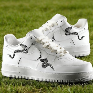 May include: White sneakers with black snake illustrations. The shoes feature a classic design with the Nike swoosh logo and a low-top silhouette. The snake graphics are detailed and add a unique touch to the footwear.