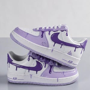 May include: White and purple custom sneakers with a dripping paint design. The sneakers feature a white base with purple accents, including the Nike swoosh, laces, and a dripping paint effect. The word "AIR" is visible on the side of the sole.