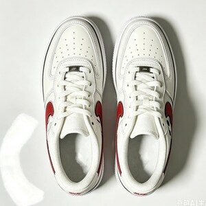 May include: Pair of white leather sneakers with red accents, including a red swoosh logo on the sides. The shoes have white laces, a white sole, and small perforations on the toe box. The design is clean and modern.