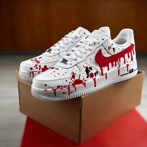Op de afbeelding: Witte sneakers met rode verfspatten en een rood swoosh-logo. De schoenen worden gepresenteerd op een bruine kartonnen doos, met het woord "AIR" aan de zijkant. Het ontwerp heeft een unieke, artistieke uitstraling.