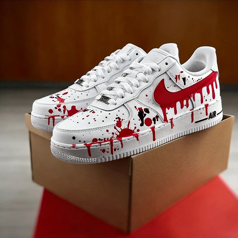 Op de afbeelding: Witte sneakers met rode verfspatten en een rood swoosh-logo. De schoenen worden gepresenteerd op een bruine kartonnen doos, met het woord "AIR" aan de zijkant. Het ontwerp heeft een unieke, artistieke uitstraling.