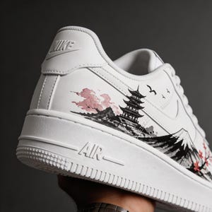 May include: White Nike Air Force 1 sneakers with a custom Japanese-inspired design. The design features a pagoda, mountains, and cherry blossoms in shades of black, pink, and red. The word "AIR" is visible on the sole.