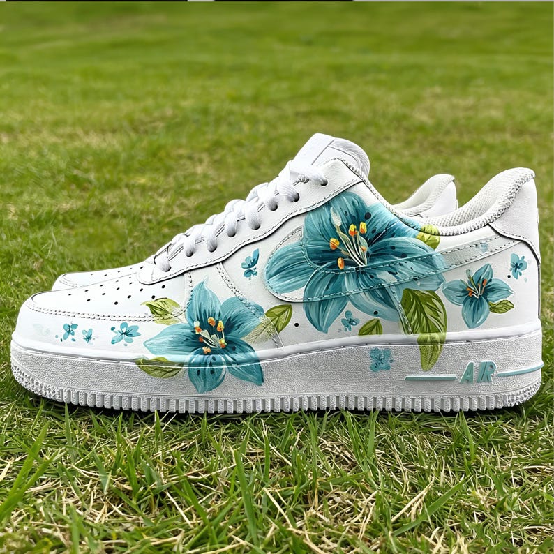 May include: White sneakers with hand-painted blue floral design. The flowers have yellow centers and green leaves. The shoes are on a green grass background.