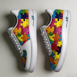 May include: Pair of white sneakers with colorful puzzle piece designs on the sides. The shoes have white laces, soles, and accents, with the letters "AR" in blue on the side.