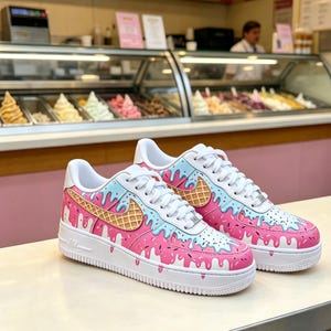May include: A pair of custom Air Force 1 sneakers with a pink, blue, and white ice cream cone design. The shoes feature a white sole, laces, and accents, with a waffle cone-shaped Nike swoosh.